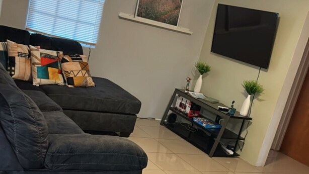 Una sala de estar con un sofá, un televisor montado en la pared, un ventilador de techo y una ventana con persianas.