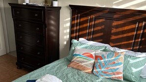 Una cama bien tendida con un edredón verde, almohadas con motivos tropicales y toallas dobladas.