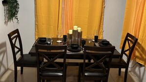 Comedor con una mesa dispuesta para cuatro personas, rodeada de sillas, un ventilador de techo y cortinas amarillas.