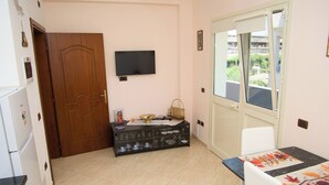 Een kamer met een houten deur, een aan de muur bevestigde televisie, een eettafel met een zwart marmeren blad en een raam met uitzicht op groen.