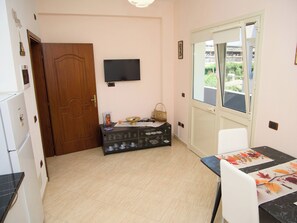Ein Zimmer mit einer Holztür, einem an der Wand befestigten Fernseher, einem Esstisch mit schwarzer Marmorplatte und einem Fenster mit Blick auf Grünfläche.
