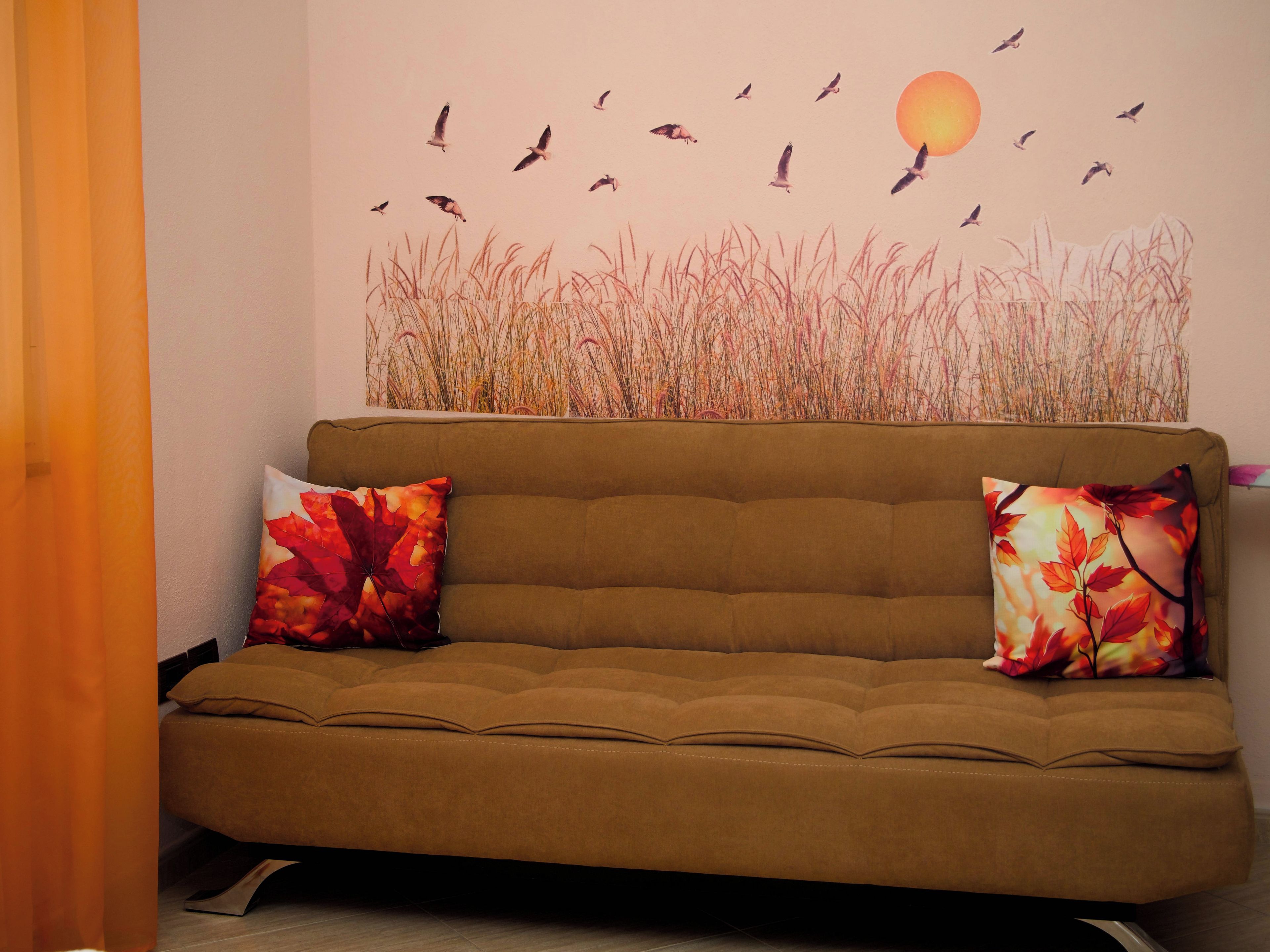 Un canapé marron avec deux coussins, une fresque murale représentant des oiseaux et un soleil, et une grande plante en arrière-plan.
