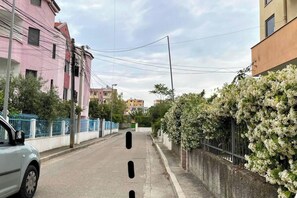 Une rue étroite bordée d’immeubles d’habitation, une voiture blanche garée sur le côté et un arbuste orné de fleurs blanches.