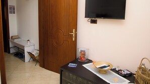 Een kamer met een houten deur, een flatscreen televisie aan de muur, een klein tafeltje met een mand, en een plank met boeken en een beker.