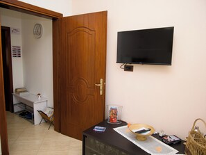 Ein Zimmer mit Holztür, einem Wand-Fernseher, einem kleinen Tisch mit einem Korb und einem Regal mit Büchern und einer Tasse.