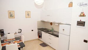 Een kleine keuken met witte kasten, een fornuis, een spoelbak en een koelkast. Twee ingelijste foto's hangen aan de muur, en op het aanrecht staan een waterkoker en een koffiezetapparaat.