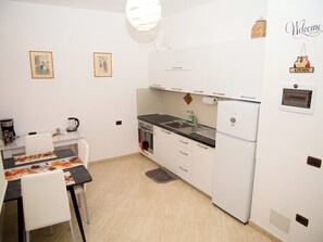 Eine kleine Küche mit weißen Schränken, einem Herd, einer Spüle und einem Kühlschrank. An der Wand hängen zwei gerahmte Bilder, auf der Arbeitsplatte stehen ein Wasserkocher und eine Kaffeemaschine.