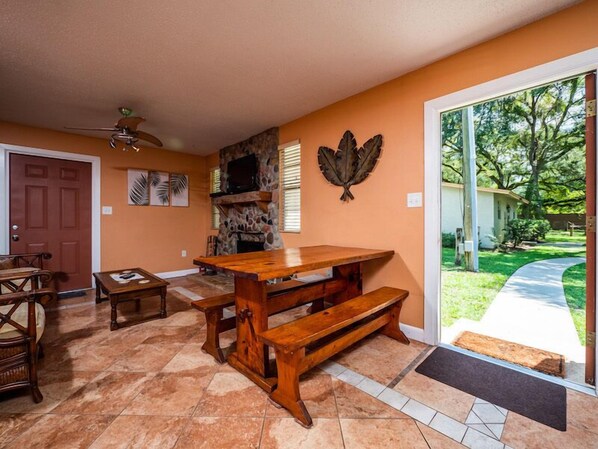 A spacious living room with a large wooden table and benches, a stone fireplace, and a view of the outdoors.