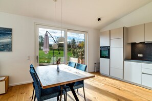 A modern kitchen with white cabinets, a wooden dining table with blue chairs, and a view of a garden through large sliding doors. - Apartment 'Winter' with Shared Pool, Private Terrace and Wi-Fi (Straubenhardt)
