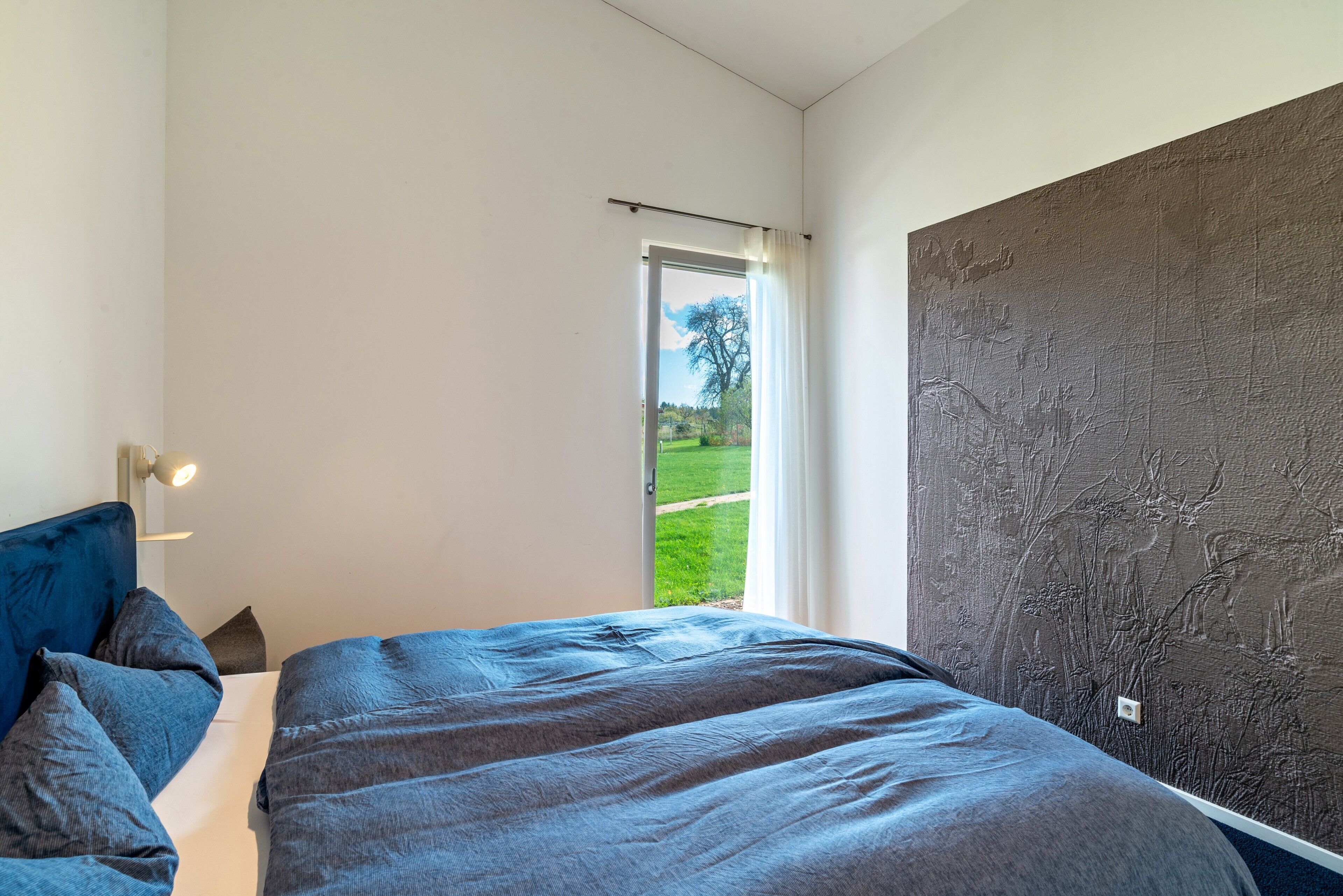 A bedroom with a bed, a nightstand, a lamp, a window with a view of a green landscape, and a textured wall.