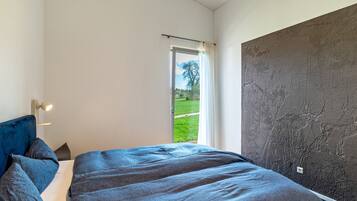 A bedroom with a bed, a nightstand, a lamp, a window with a view of a green landscape, and a textured wall.