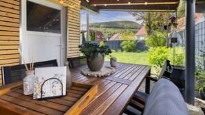 Une terrasse en bois avec une table, des chaises, un pot de fleurs et une vue sur une maison et des collines.