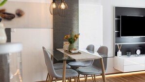 Un espace repas moderne avec une table en verre, des chaises grises, un vase de fleurs et un téléviseur fixé au mur.