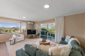 A spacious living room with a large flat-screen TV, a sofa, a coffee table, and a view of a garden through large windows. - Kinder Cottage (Taupo)