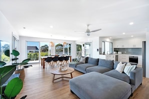 A modern living space with a dining area, a grey sofa, and a kitchen with white cabinets. - Palm Stays | Ocean views | Space & style | Large family beach house (Vincentia)