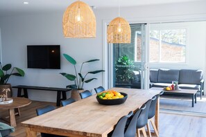 A modern dining area with a wooden table, black chairs, a bowl of fruit, and two hanging rattan lights. - Palm Stays | Ocean views | Space & style | Large family beach house (Vincentia)