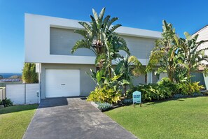 A modern two-story house with a white garage door, a concrete driveway, and lush tropical landscaping. - Palm Stays | Ocean views | Space & style | Large family beach house (Vincentia)