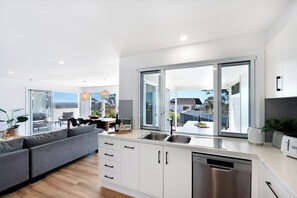 A modern kitchen with white cabinetry, a stainless steel dishwasher, and a large window offering a view of the outdoors. - Palm Stays | Ocean views | Space & style | Large family beach house (Vincentia)