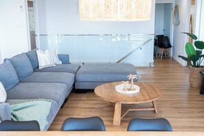 A modern living room with a sectional sofa, a round wooden coffee table, and a glass railing in the background. - Palm Stays | Ocean views | Space & style | Large family beach house (Vincentia)