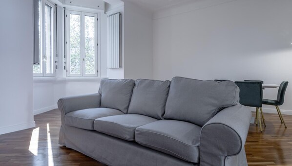 A grey sofa in a room with wooden flooring, white walls, and a window with white shutters.