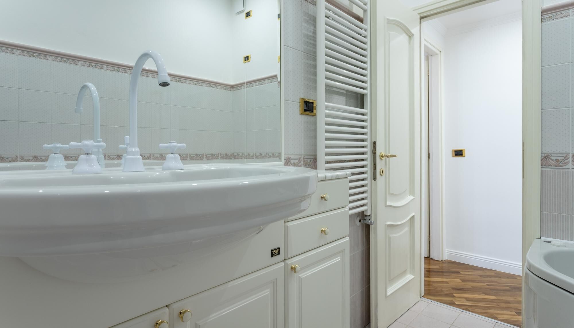 Une salle de bain avec un grand lavabo blanc, plusieurs robinets, un miroir et une porte donnant sur une autre pièce.