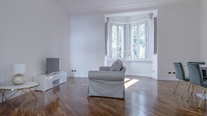 A spacious living room with a grey sofa, a small white side table with a lamp, a flat-screen TV, and a dining area with two chairs. - Italianway Comfort - Bodio 6 (Milan)