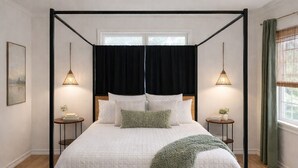 A four-poster bed with a white quilt, two bedside tables with lamps, a green throw blanket, and a patterned rug.