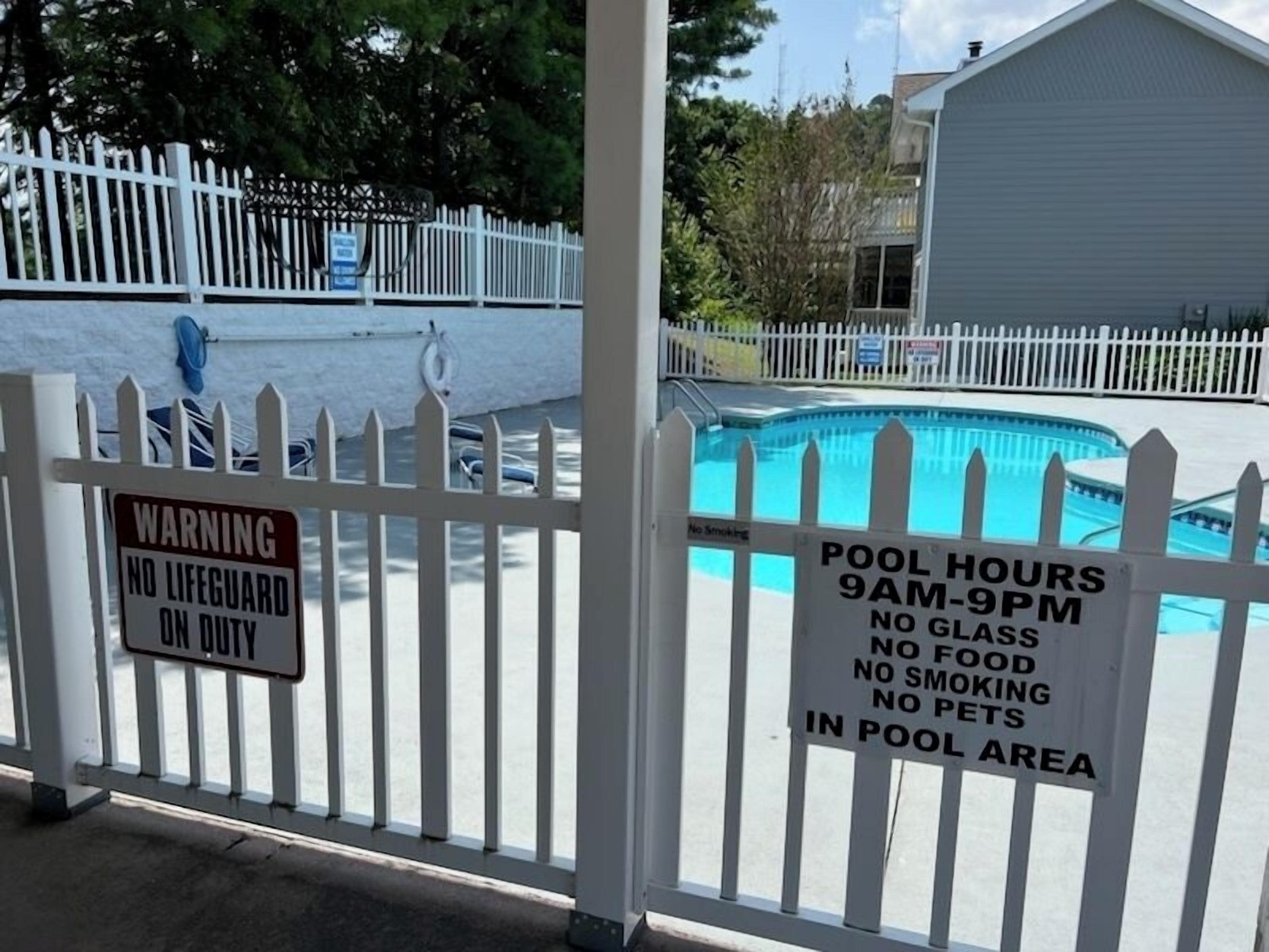 Zona de piscina con cerca blanca, aviso indicando ausencia de socorrista, y señal con el horario de la piscina, prohibido fumar, comer o mascotas.