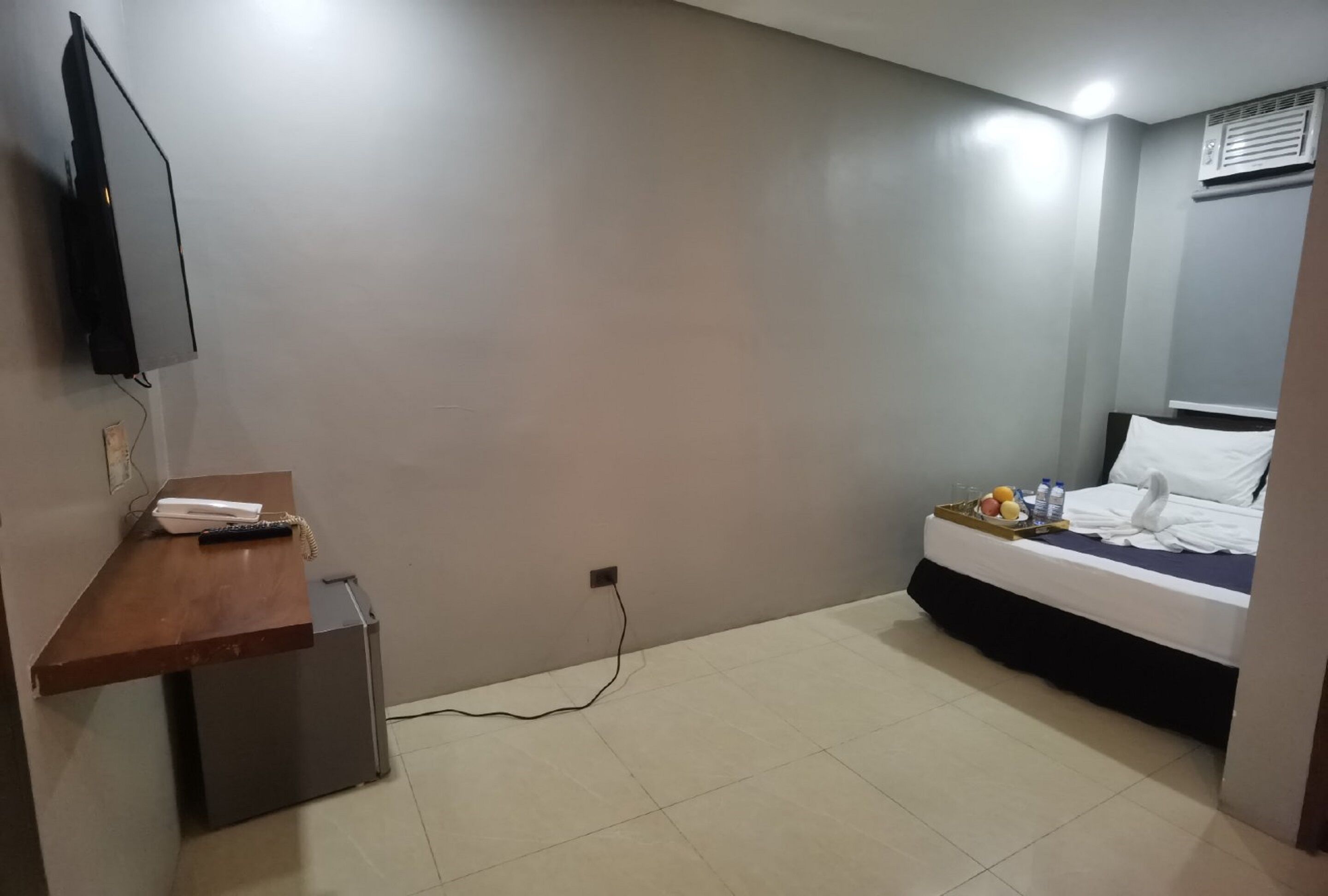 A hotel room with a bed, a desk with a phone, a small refrigerator, and a TV mounted on the wall.