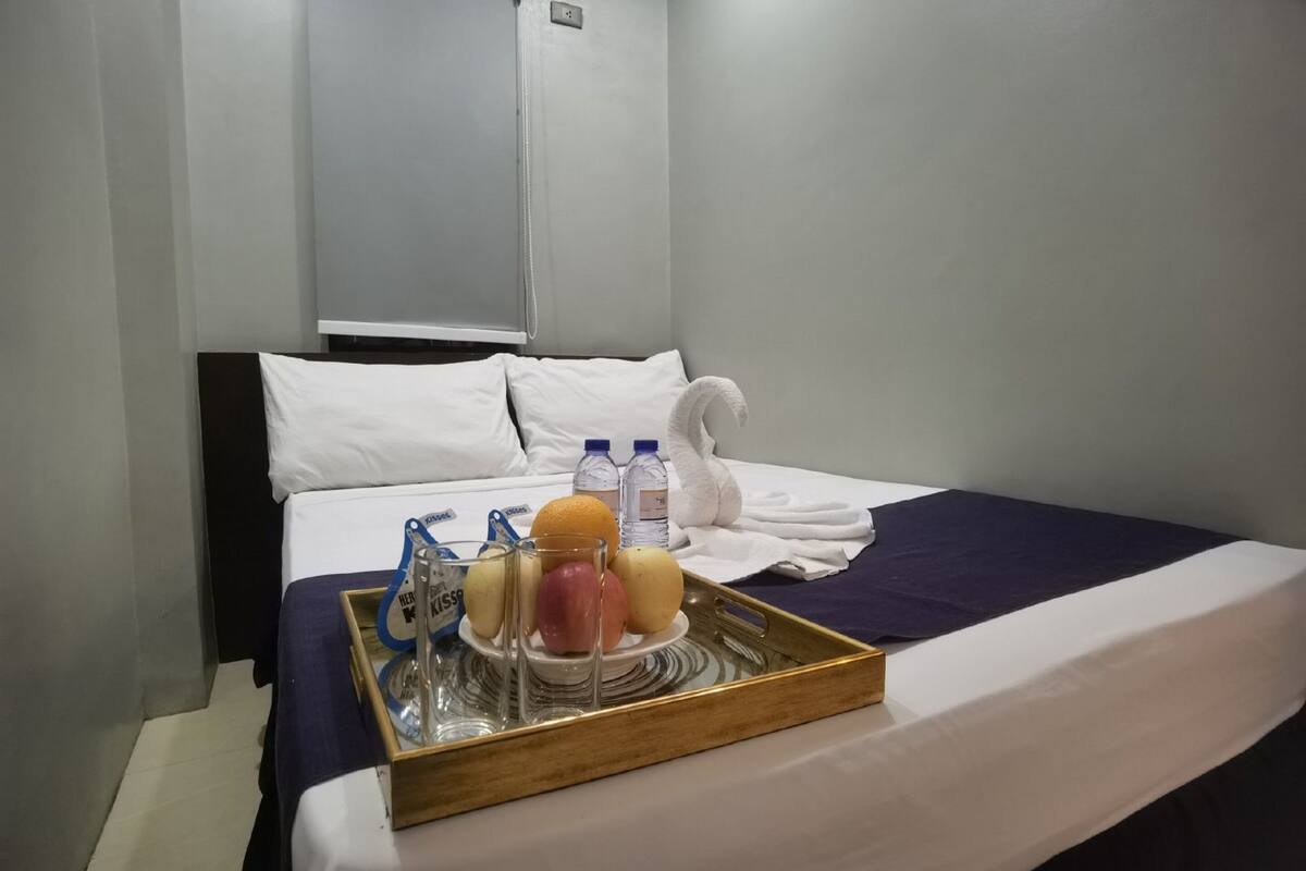 A hotel room with a neatly made bed, a tray with fruits and bottled water, and a towel on the bed.