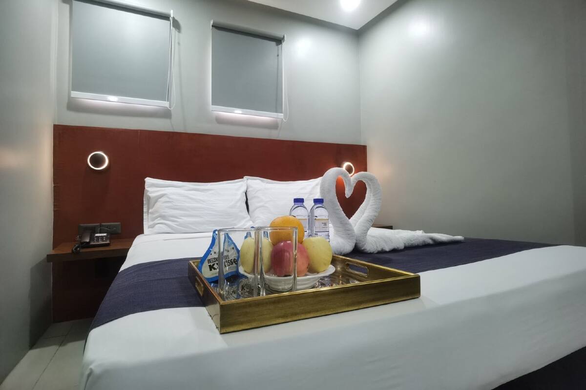 A hotel room with a neatly made bed, a tray with fruits and a bottle, and a heart-shaped towel on the bed.