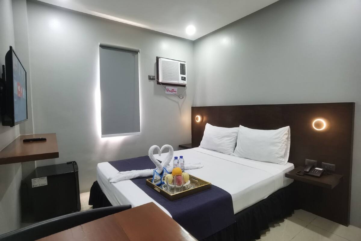 A hotel room with a bed, bedside table, nightstand, and a wall-mounted air conditioner.