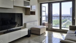 A modern living room with a flat-screen TV, a sofa, and a large sliding glass door leading to a balcony with a city view.