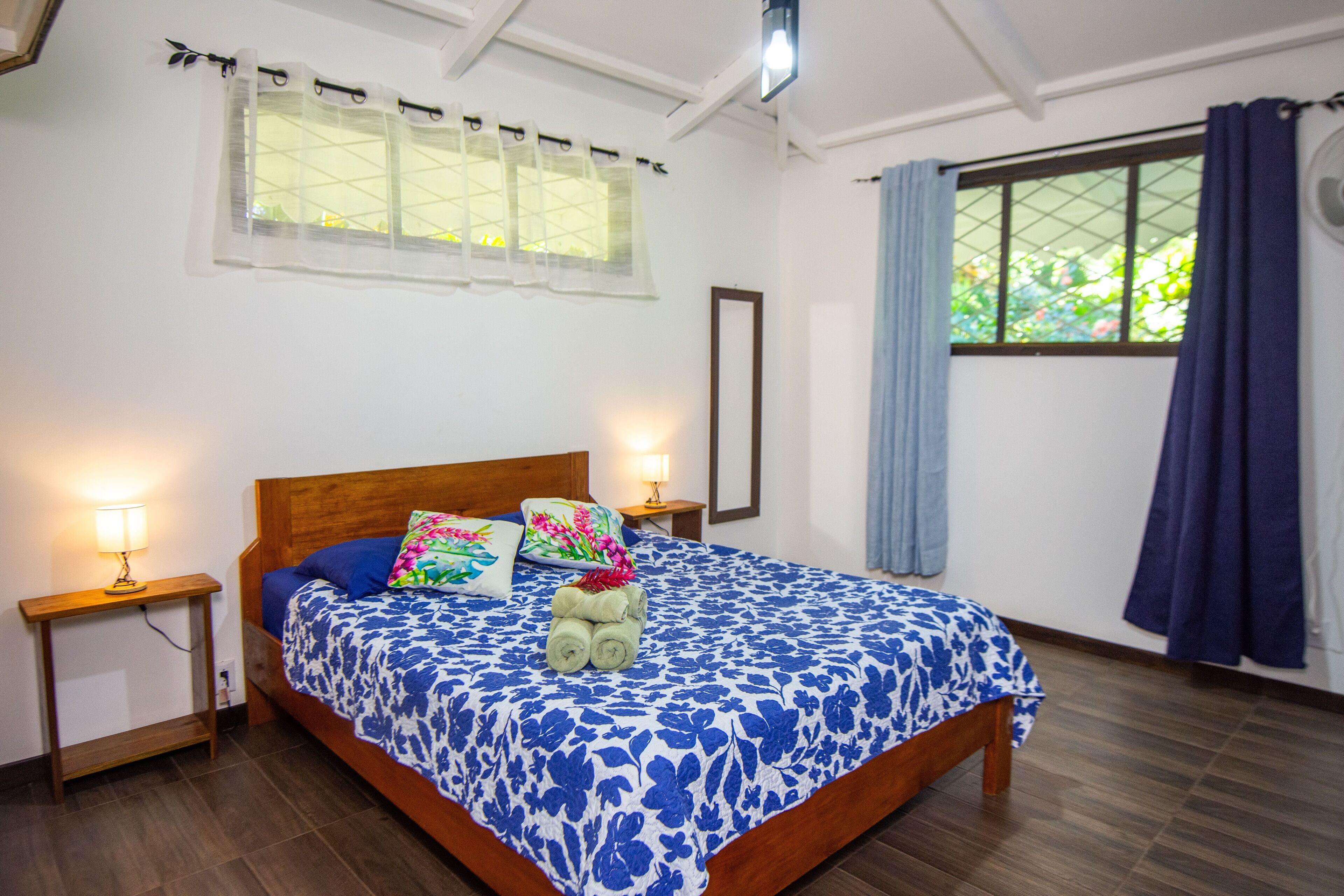 Una cama bien hecha con una colcha estampada con motivos florales, dos lámparas de noche y una ventana con vistas a la vegetación.