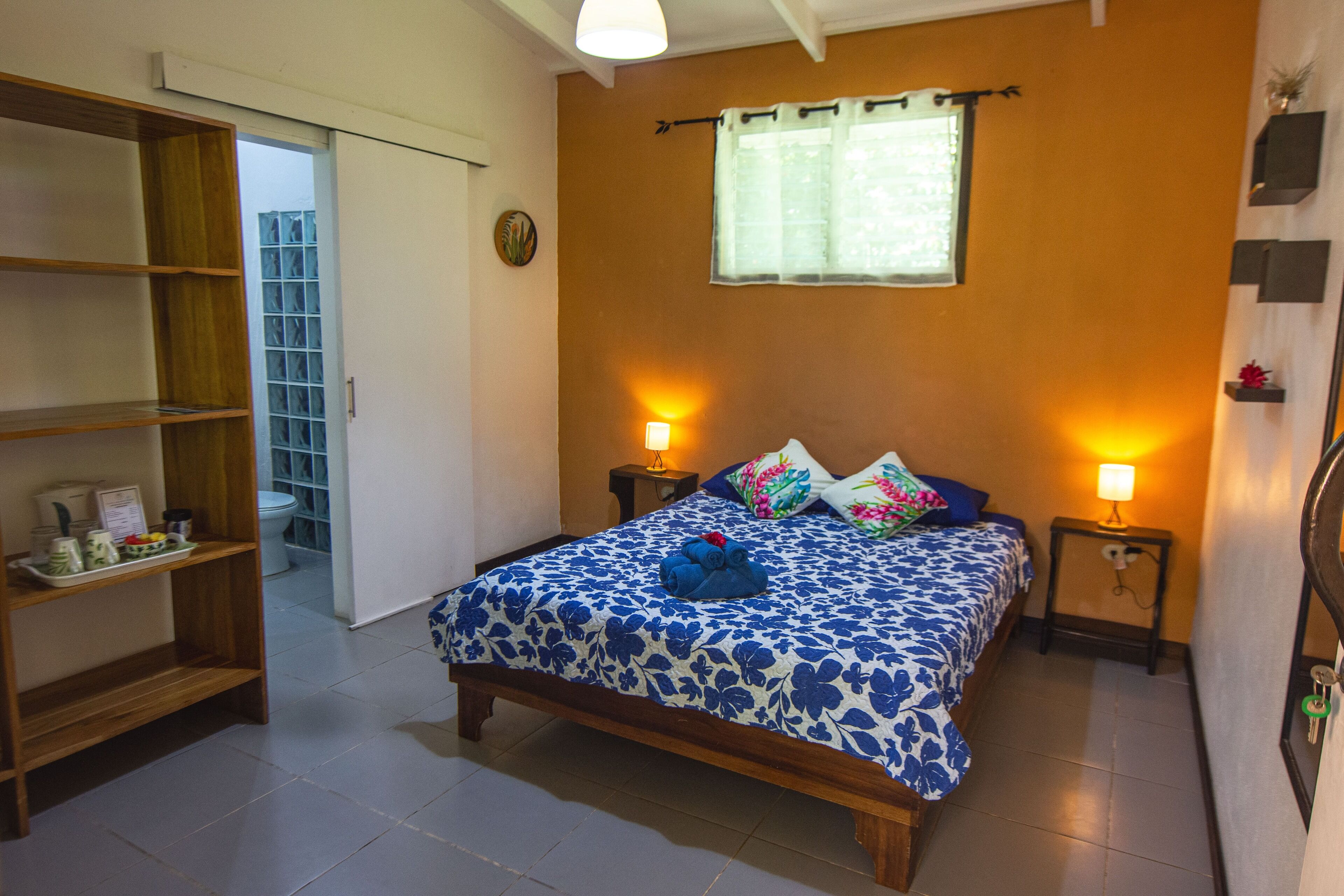 A bedroom with a bed, two bedside tables, a wooden shelf unit, a window with curtains, and a door leading to a bathroom.