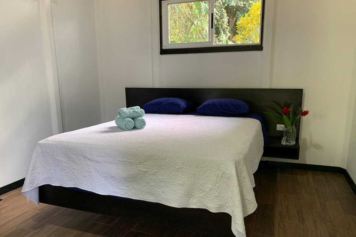 A neatly made bed with a white textured bedspread, a dark headboard, and two blue pillows. A small stack of folded towels is placed on the bed, and a vase with red flowers is on a nightstand beside the bed.