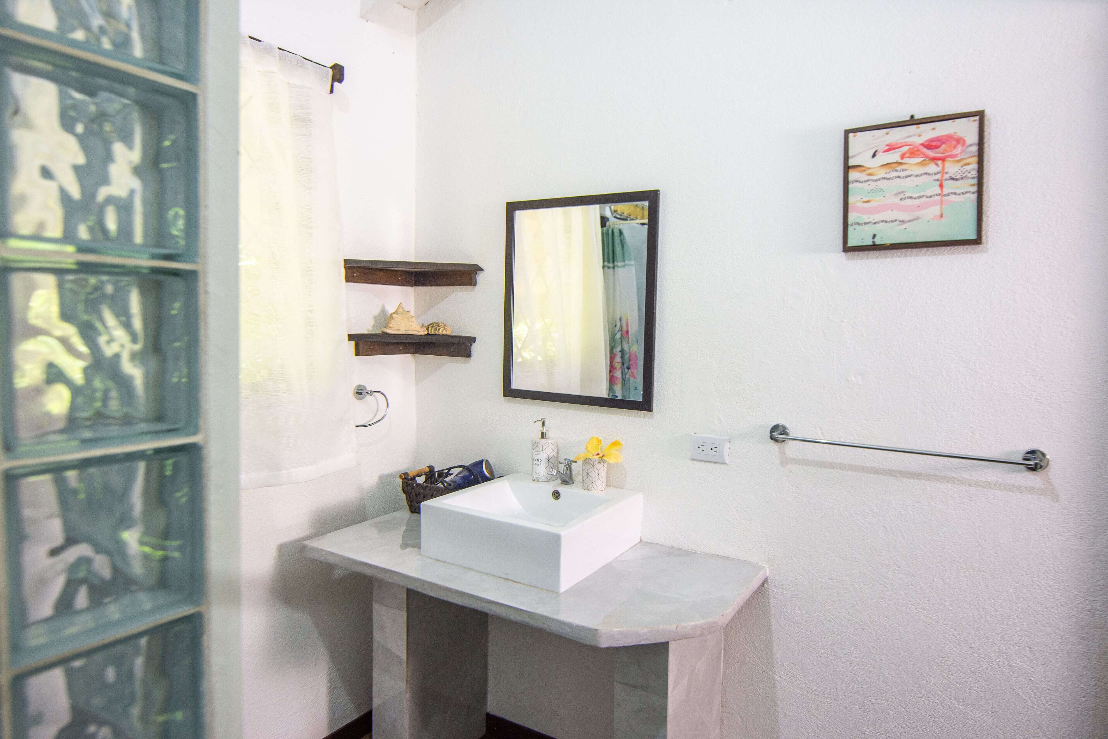 Un baño con lavamanos blanco, un espejo, una obra de arte enmarcada y un perchero para toallas.