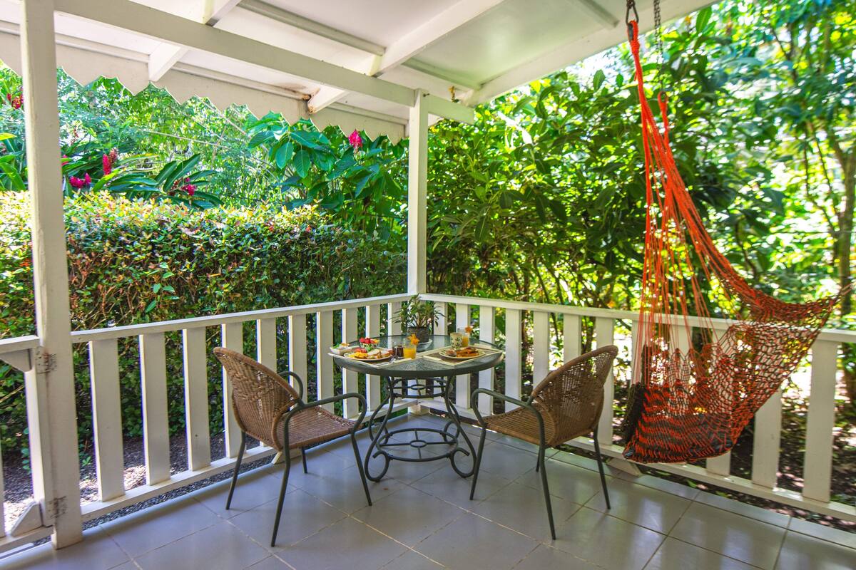 A covered patio with a table and chairs, surrounded by lush greenery and a hanging hammock.