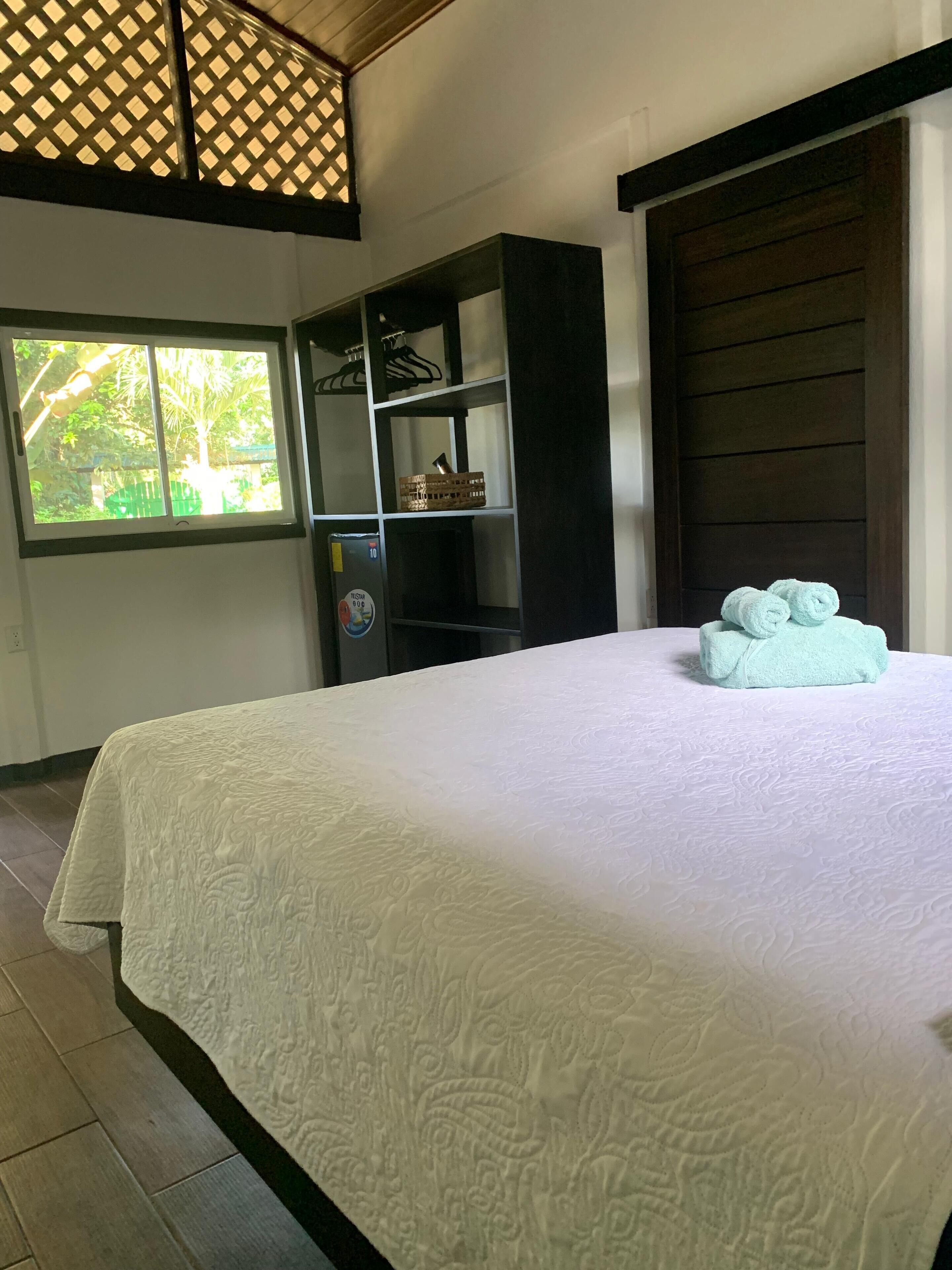 A neatly made bed with a white bedspread and two rolled towels. There is a wooden wardrobe with a mirror and a shelf with a basket. A window with a view of greenery is visible.