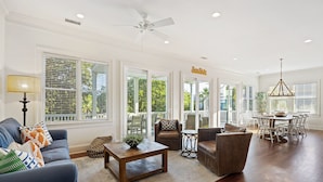 A spacious living room with a ceiling fan, large windows, and a dining area with a chandelier.