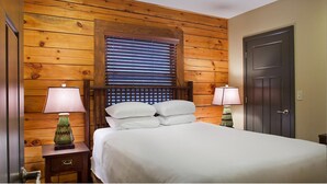 Une chambre avec un mur lambrissé en bois, une fenêtre avec des stores, un lit recouvert de draps blancs, deux tables de chevet avec des lampes et une porte.