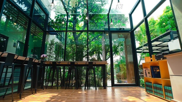 A modern interior with a wooden table and chairs, a glass wall with a view of greenery, and a bookshelf with books. - Bed Garden 26 (Bangkok)