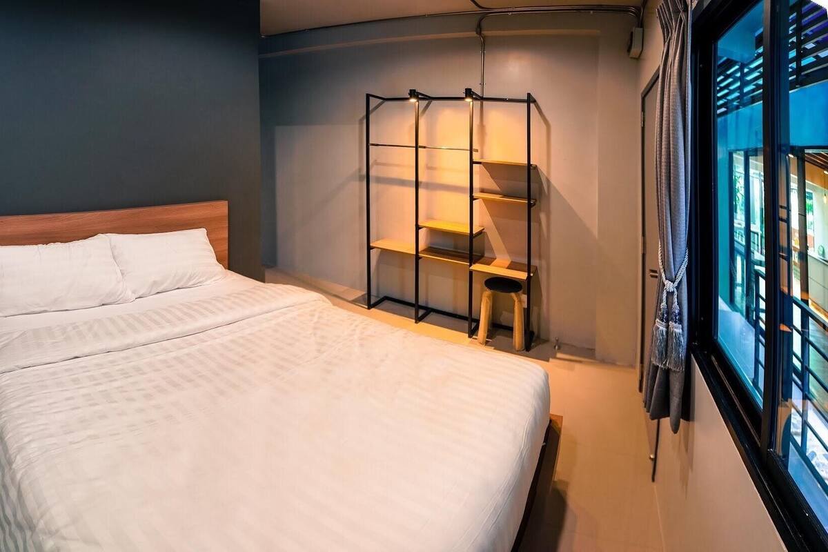 A bed with a wooden headboard, a small wooden stool, and a metal shelf unit with a lamp.