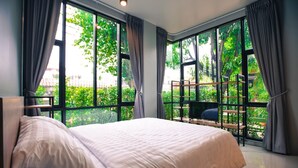 A bedroom with a large bed, a view of greenery through large windows, and a balcony with a seating area. - Bed Garden 26 (Bangkok)