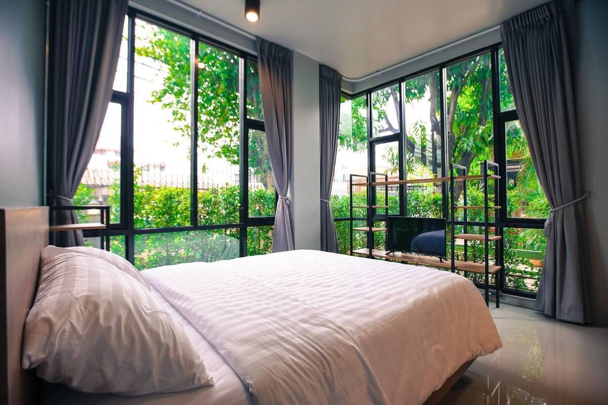 A bedroom with a large bed, a view of greenery through large windows, and a balcony with a seating area.