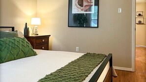 Une chambre à coucher avec un lit, une table de chevet, une lampe, un tableau encadré au mur et une porte donnant sur une autre pièce.