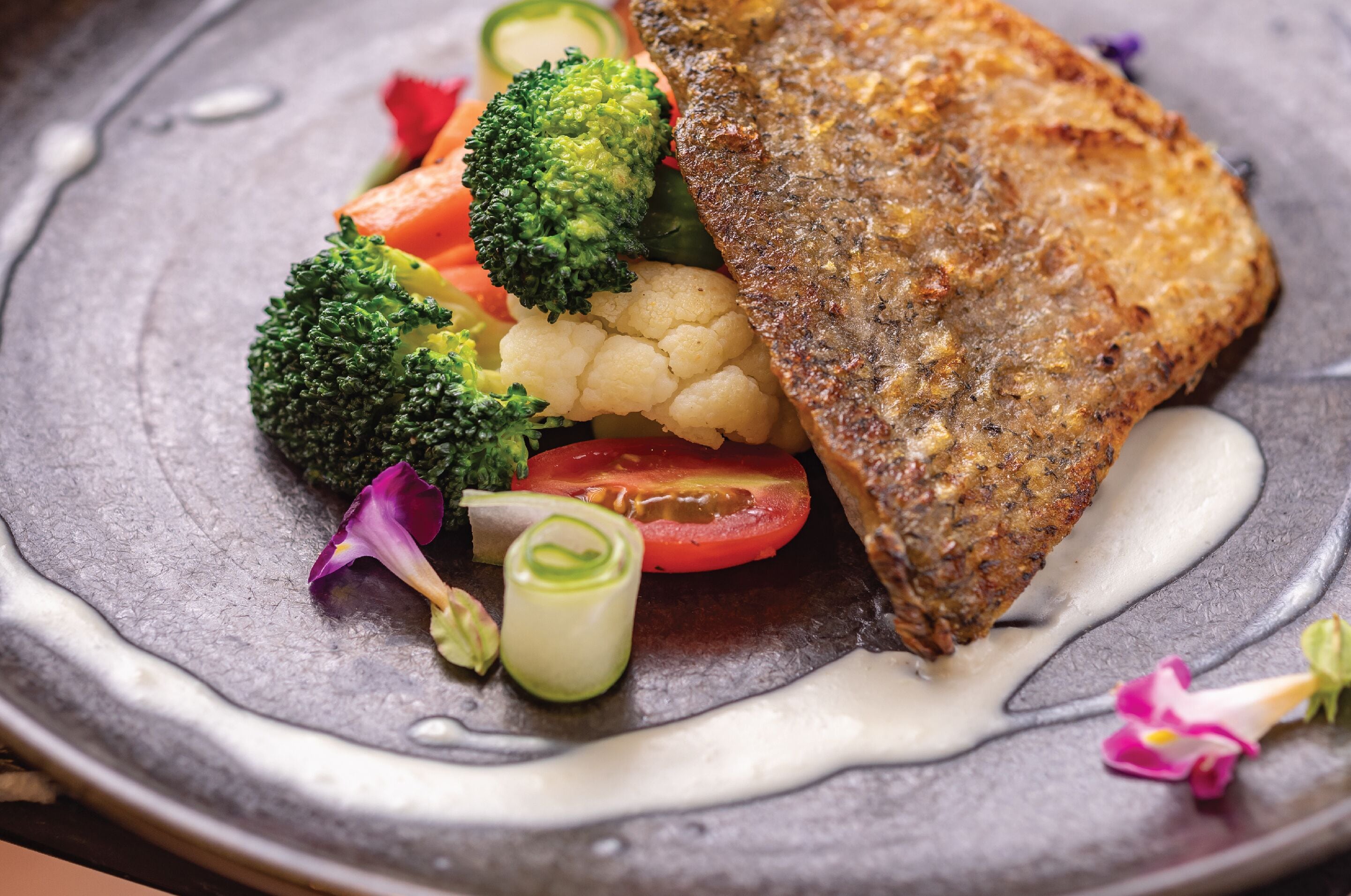 A plate with a piece of fried fish, broccoli, cauliflower, and tomato slices, garnished with a creamy sauce and edible flowers.