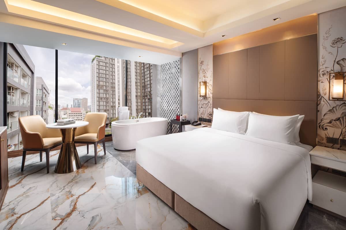 A modern hotel room with a large bed, a round table, two armchairs, a bathtub, and a city view.