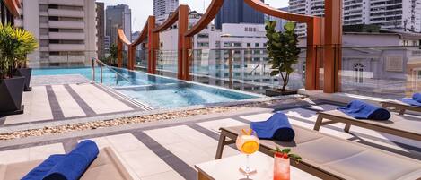 Zona de piscina en la azotea con sillones reclinables, una mesa con bebidas y vistas a edificios de la ciudad.