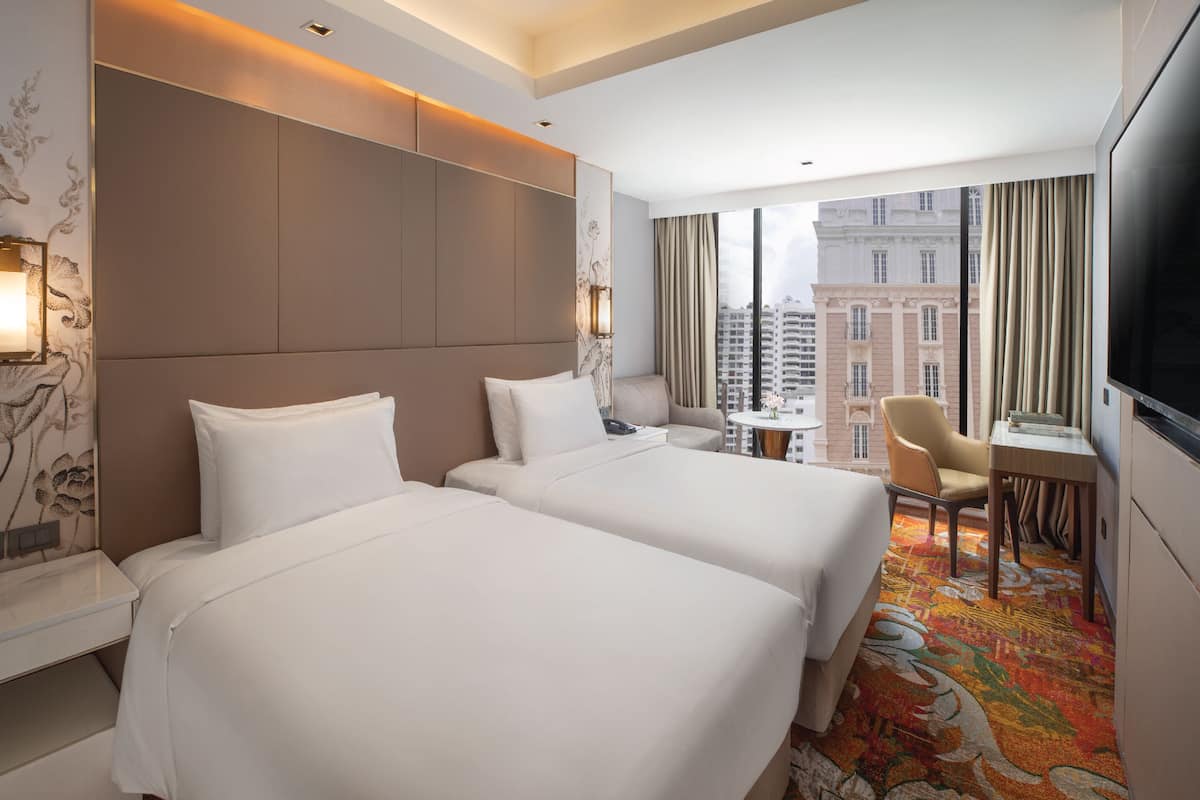 A hotel room with two beds, a desk, a chair, a TV, and a view of a cityscape.